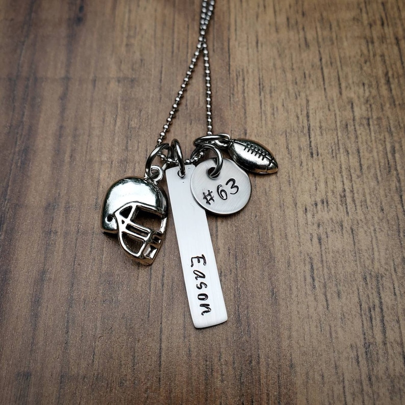May include: A silver-toned necklace featuring a ball chain and three charms: a football helmet, a rectangular bar pendant engraved with the name Eason, and a circular disc pendant stamped with the text #63, alongside a small football charm.