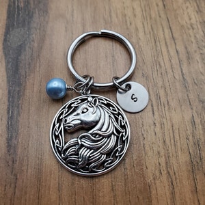 May include: Silver-toned horse keychain with a round pendant featuring a horse head design within a Celtic knot border. Includes a small round charm with the letter "S" and a blue bead. The keychain is on a silver ring.