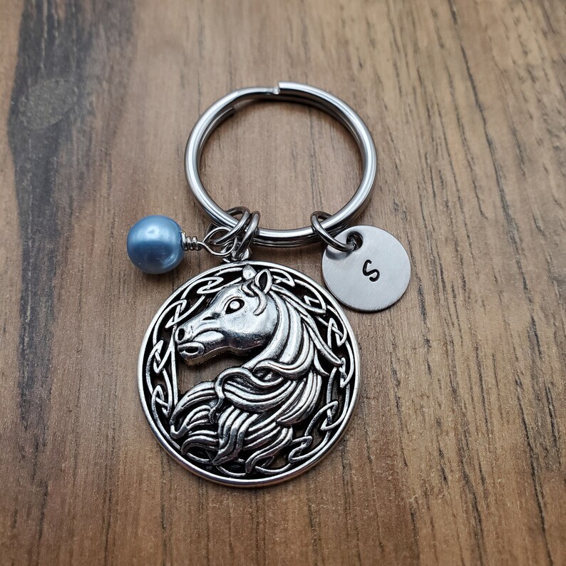 May include: Silver-toned horse keychain with a round pendant featuring a horse head design within a Celtic knot border. Includes a small round charm with the letter "S" and a blue bead. The keychain is on a silver ring.