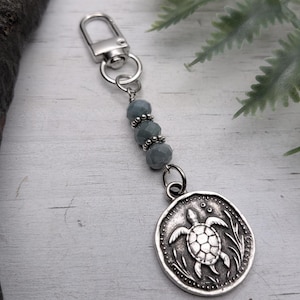 Peut inclure: Porte-clés argenté avec un pendentif rond représentant une tortue. Le pendentif est relié à une chaîne de perles grises et à un fermoir argenté. Le fond est une surface en bois clair avec des feuilles vertes.