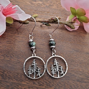 May include: Silver-toned dangle earrings featuring a circular design with a tree silhouette. Each earring has a green and silver bead above the circular pendant. The earrings are displayed on a wooden surface with pink flowers in the background.