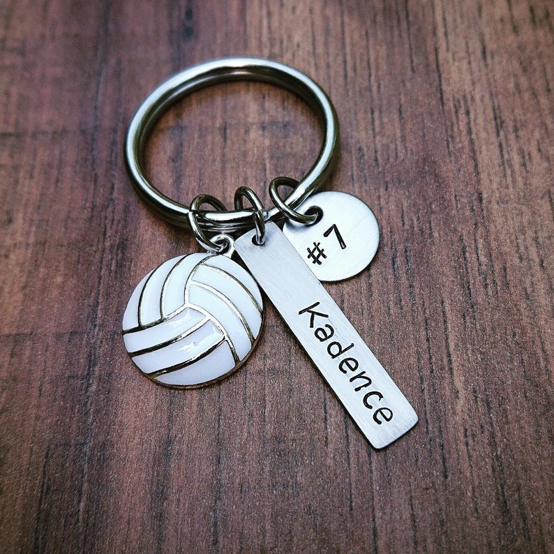 May include: Silver volleyball keychain with a white volleyball charm, a silver charm with the number 7, and a silver tag with the name Kadence.