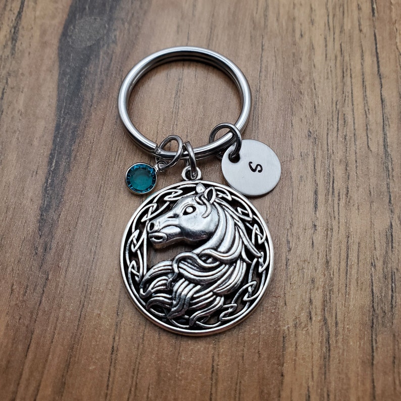 May include: Silver-toned horse keychain with a teal-colored charm and a small round tag with the letter "S". The horse pendant is circular with a Celtic knot design around the horse's head. The keychain is on a wooden surface.
