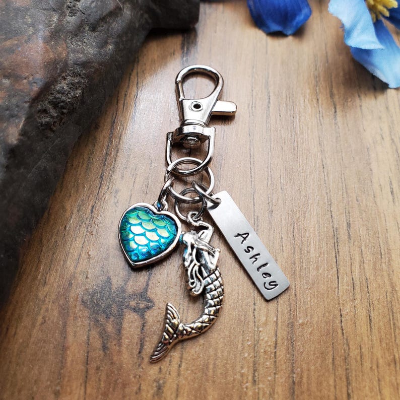 Puede incluir: Un llavero plateado con cierre de mosquet&oacute;n, con un dije de sirena, un dije en forma de coraz&oacute;n con dise&ntilde;o de escamas turquesas y una etiqueta rectangular grabada con el nombre "Ashley".