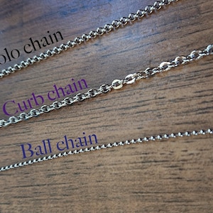 May include: Three different types of silver chains: rolo chain, curb chain, and ball chain. The chains are laid out on a wooden surface.
