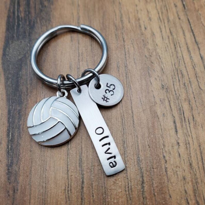 May include: Silver volleyball keychain with a white volleyball charm, a silver bar charm with the name "Olivia" engraved on it, and a silver round charm with the number "#35" engraved on it.