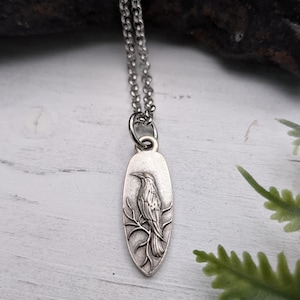 May include: Silver-toned pendant necklace featuring a detailed raven perched on a branch. The oval pendant hangs from a delicate chain. The raven is intricately designed, showcasing feathers and a detailed beak. The necklace is displayed against a rustic background.
