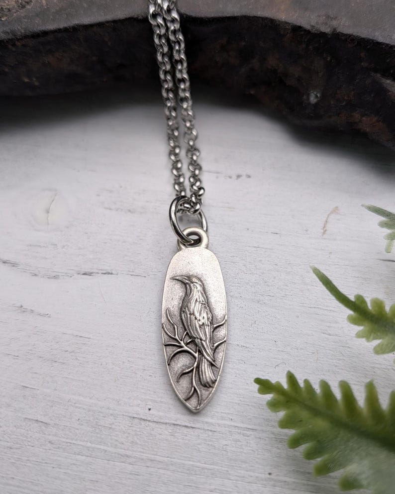 May include: Silver-toned pendant necklace featuring a detailed raven perched on a branch. The oval pendant hangs from a delicate chain. The raven is intricately designed, showcasing feathers and a detailed beak. The necklace is displayed against a rustic background.