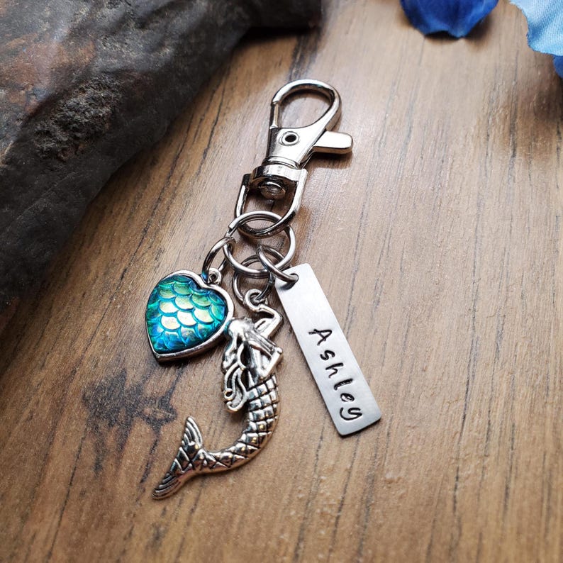 Puede incluir: Un llavero plateado con cierre de mosquet&oacute;n. Cuenta con un dije de sirena, un dije en forma de coraz&oacute;n con escamas azules y verdes, y una etiqueta grabada con el nombre "Ashley".