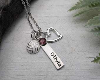 May include: A silver-toned necklace featuring a vertical rectangular pendant engraved with the name Olivia, a small round dark red gemstone, a volleyball charm, and an open heart charm, all suspended from a delicate silver-toned chain.