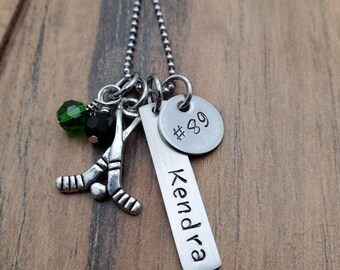 May include: Silver necklace with a hockey stick charm, a green and black crystal charm, and a silver tag with the name "Kendra" and the number "#89" engraved on it.