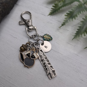 May include: Silver keychain with a lobster clasp, featuring charms of a raccoon, acorn, leaf, a small disc with the letter "B", and a rectangular pine tree design. The raccoon charm is black, white, and grey. The leaf charm is green and gold.
