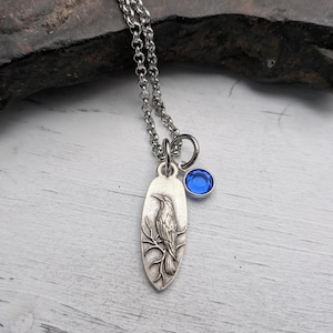 May include: Silver-toned pendant necklace featuring a detailed raven design on an oval pendant. A small, round, blue gemstone charm hangs next to the pendant. The necklace has a delicate chain.