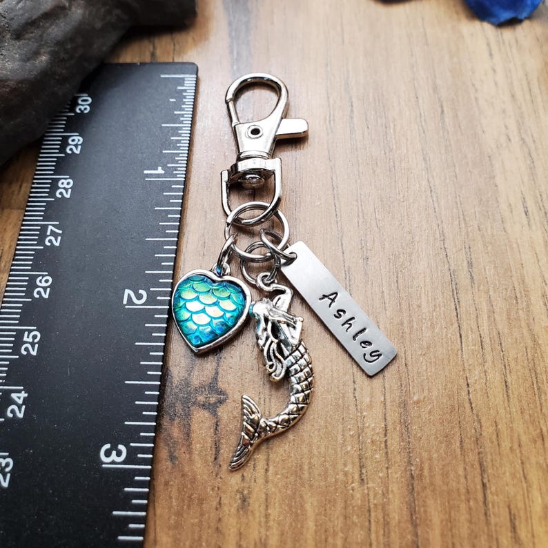 Puede incluir: Un llavero plateado con un dije de sirena, un dije en forma de coraz&oacute;n con un patr&oacute;n de escamas azul y verde y una etiqueta con el nombre "Ashley". El llavero incluye un mosquet&oacute;n. Una regla muestra las medidas en cent&iacute;metros.