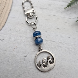 May include: Silver-coloured keychain with a lobster clasp, blue beads, and a circular charm with a wave design. The keychain is attached to a silver ring, likely made of metal.