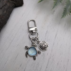 Peut inclure: Porte-clés argenté avec un charm en forme de tortue de mer avec un centre bleu clair et un charm en forme de dollar des sables. Le porte-clés a un fermoir mousqueton. Les breloques sont détaillées et sur le thème de l'océan.