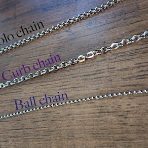May include: Close-up of three silver chain styles: Rolo chain, Curb chain, and Ball chain. The chains are displayed on a wooden surface. Text labels identify each chain type. The Rolo chain has oval links, the Curb chain has flattened links, and the Ball chain has small spheres.
