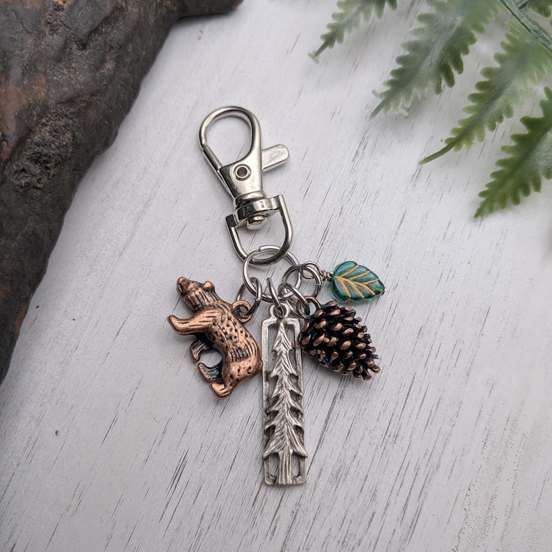 May include: A silver-tone keychain with a lobster clasp, featuring charms of a bear, a pine tree, a pine cone, and a green and gold leaf. The bear charm is bronze-colored. The pine cone is brown.