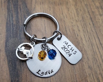 Puede incluir: Un llavero de plata con un dije redondo que dice "Lydia" y un dije más pequeño con un gorro de graduación y el texto "MCHS 2024".