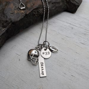 May include: A silver-toned ball chain necklace featuring three charms: a miniature football helmet, a round disc engraved with #33, and a rectangular vertical bar tag engraved with the name Bishop, alongside a small football charm.