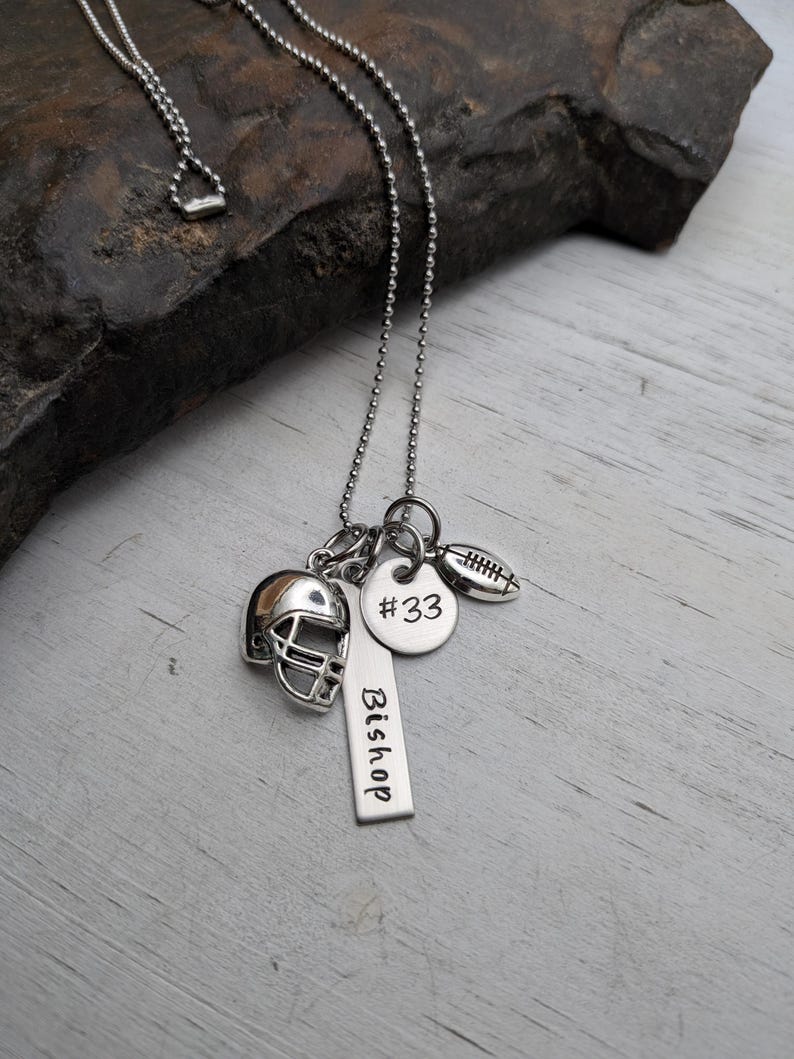 May include: A silver-toned ball chain necklace featuring three charms: a miniature football helmet, a round disc engraved with #33, and a rectangular vertical bar tag engraved with the name Bishop, alongside a small football charm.