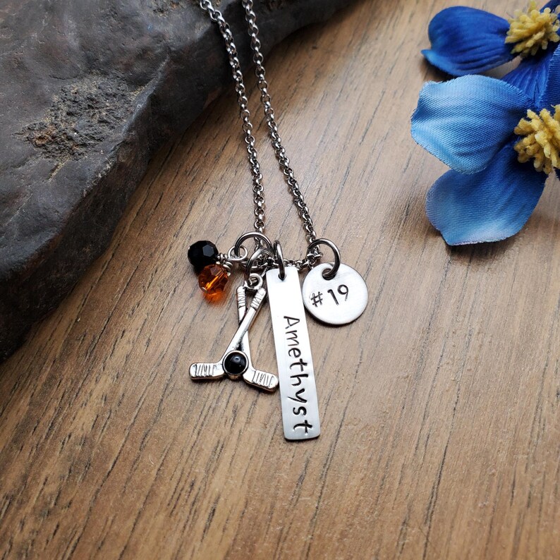 May include: Silver necklace with a hockey stick charm, a round charm with the number 19, and a bar charm with the word "Amethyst".