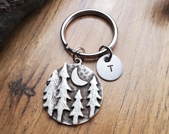 May include: Silver keychain with a charm featuring a crescent moon and five pine trees. The charm is attached to a silver key ring and a silver disc with the letter "T" engraved on it.