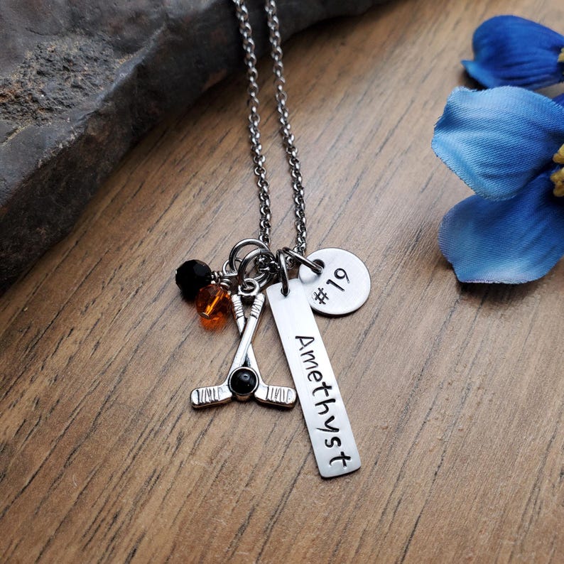 May include: A silver necklace with a hockey stick charm, a black bead, orange beads, and a silver tag that says "Amethyst" and a silver disc that says "#19".