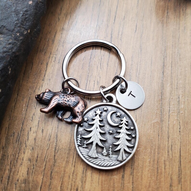 May include: A silver keychain with a bear charm and a round pendant featuring trees, a crescent moon, and stars. Includes a small, round charm with the letter "T". The bear charm is brown and copper-colored.