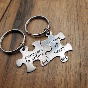 May include: Two silver puzzle piece keychains with the text "partners in crime" and "sisters at heart" on them. The keychains are connected together and have the initials "R&L" on them.
