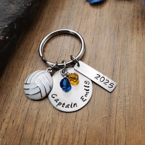 May include: A silver keychain with a volleyball charm, a round charm that says "Captain", a charm with blue and yellow beads, and a rectangular charm that says "Emily 2025".
