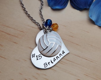 May include: Silver-toned necklace with a heart-shaped pendant. The pendant features a white volleyball design and the name "Brianna" with the number "15". The necklace has a chain and is adorned with blue and orange beads.