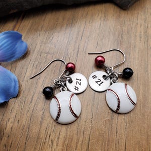 May include: A pair of silver earrings with a baseball charm and a silver disc with the number 21 on it. The earrings have red and black beads.