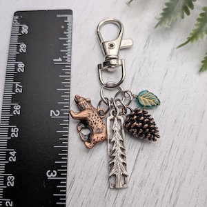 May include: A silver-tone keychain with a lobster clasp, featuring charms of a copper-colored bear, a silver tree, a pinecone, and a teal leaf. The keychain is approximately 2.5 inches long, perfect for keys or a bag.