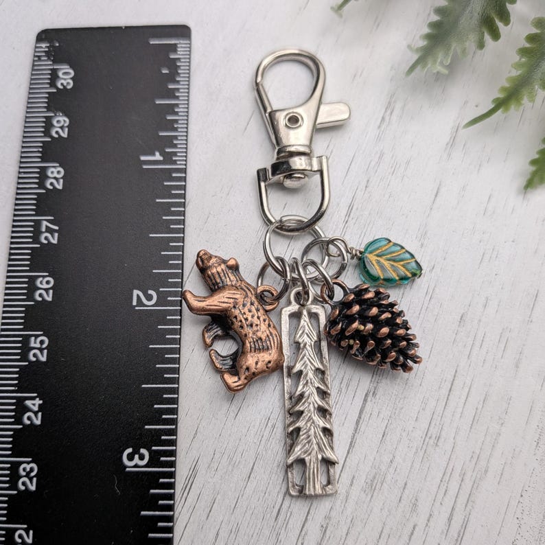 May include: A silver-tone keychain with a lobster clasp, featuring charms of a copper-colored bear, a silver tree, a pinecone, and a teal leaf. The keychain is approximately 2.5 inches long, perfect for keys or a bag.