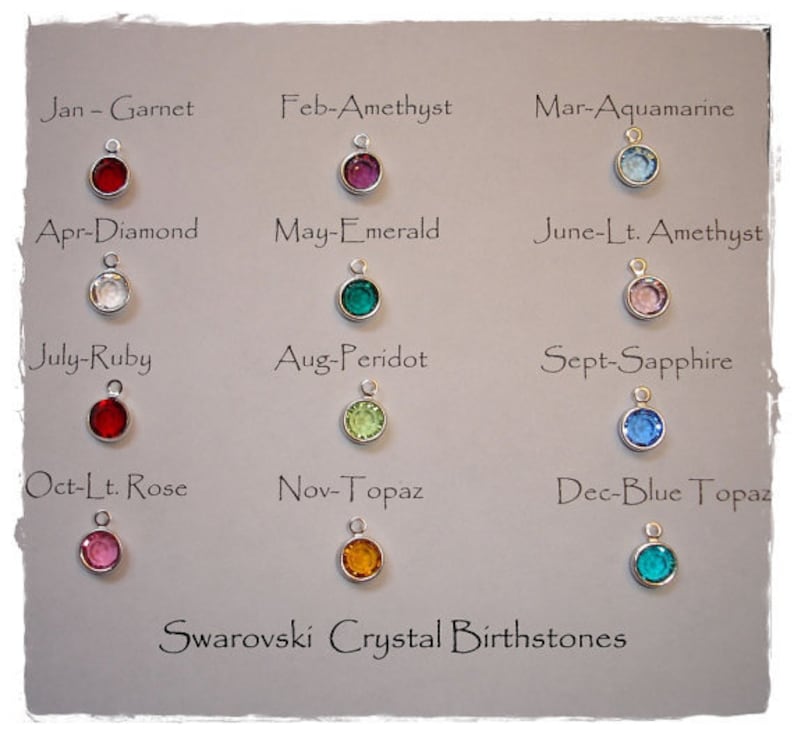 Puede incluir: Una exhibici&oacute;n de dijes de piedras de nacimiento de cristal Swarovski en varios colores, incluyendo rojo, morado, verde y azul. Cada dije est&aacute; etiquetado con un mes y el nombre de una gema, como "Ene - Granate". El texto "Swarovski Crystal Birthstones" est&aacute; en la parte inferior.