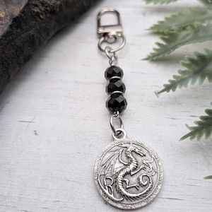 May include: A silver-toned metal keychain featuring a circular pendant with an embossed dragon design. The pendant is attached to a silver lobster clasp by a short chain containing three faceted, round, black beads arranged in a vertical line.