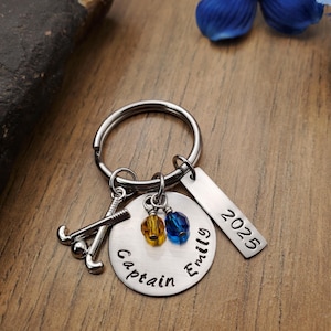 May include: A silver keychain with a hockey stick charm, a round charm that says "Captain", a charm with the name "Emily", and a charm with the year "2025". The charms are attached to a silver key ring with blue and yellow beads.
