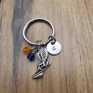 May include: Silver keychain with a winged shoe charm, blue and orange beads, and a silver disc with the letter 'G' engraved on it.