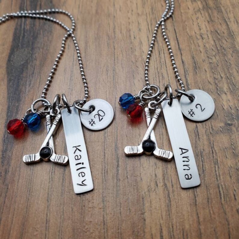 May include: Two silver necklaces with hockey stick charms, red and blue crystal beads, and personalized tags. One tag reads "Kailey #20" and the other reads "Anna #2".