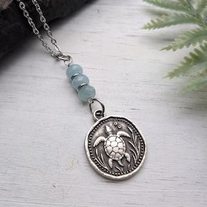 Peut inclure: Un collier argenté avec un pendentif rond représentant une tortue. Le pendentif est relié à une chaîne ornée de trois perles bleu clair. Le collier est présenté sur une surface en bois blanc.