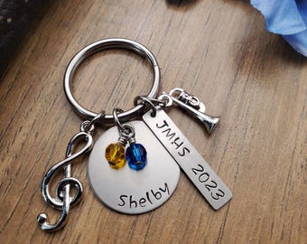 May include: A silver keychain with a music note charm, a round charm with the name "Shelby" engraved on it, a charm with the text "JMHS 2023" and a trumpet charm. The charms are attached to a silver key ring.