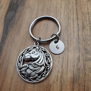 May include: Silver-toned horse head keychain with a Celtic knot design around the horse's head. The keychain includes a small, round charm with the letter "S" and a silver key ring. The background is a wooden surface.