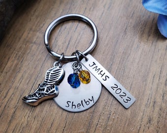 May include: A silver keychain with a running shoe charm, two blue and yellow beads, a round tag that says "Shelby", and a rectangular tag that says "JMHS 2023".