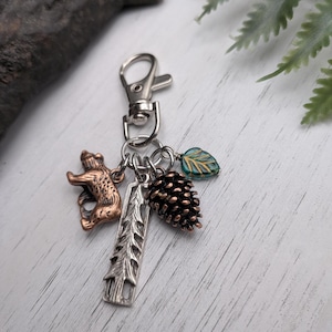 May include: A silver-toned keychain with a lobster clasp, featuring charms of a bear, pine cone, leaf, and a tree. The bear is bronze-colored, the pine cone is dark brown, and the leaf is teal and gold. The tree charm is silver.