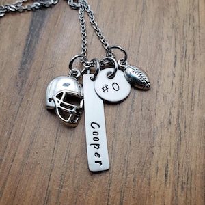 May include: A silver-toned necklace featuring three charms: a football helmet, a rectangular bar pendant engraved with the name Cooper, and a circular disc pendant stamped with the text #0, accompanied by a small football-shaped charm.