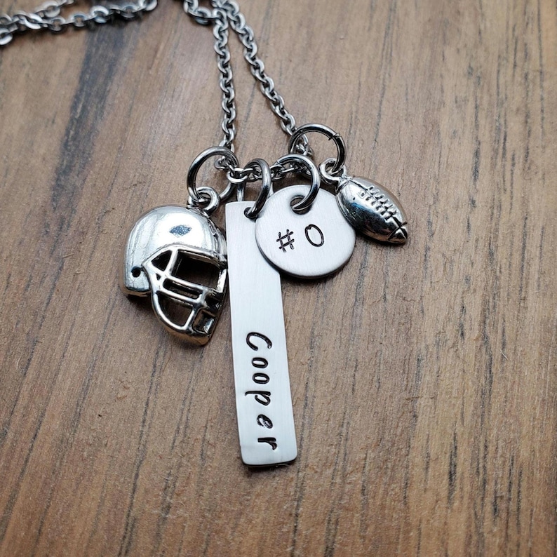 May include: A silver-toned necklace featuring three charms: a football helmet, a rectangular bar pendant engraved with the name Cooper, and a circular disc pendant stamped with the text #0, accompanied by a small football-shaped charm.