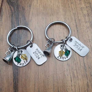 May include: Two silver keychains with charms. Each keychain has a silver tag with the text "WMVC 2024". One keychain has a charm with the name "Tayla" and the other has a charm with the name "Lillie". Both keychains have a silver whistle charm and green and yellow crystal charms.