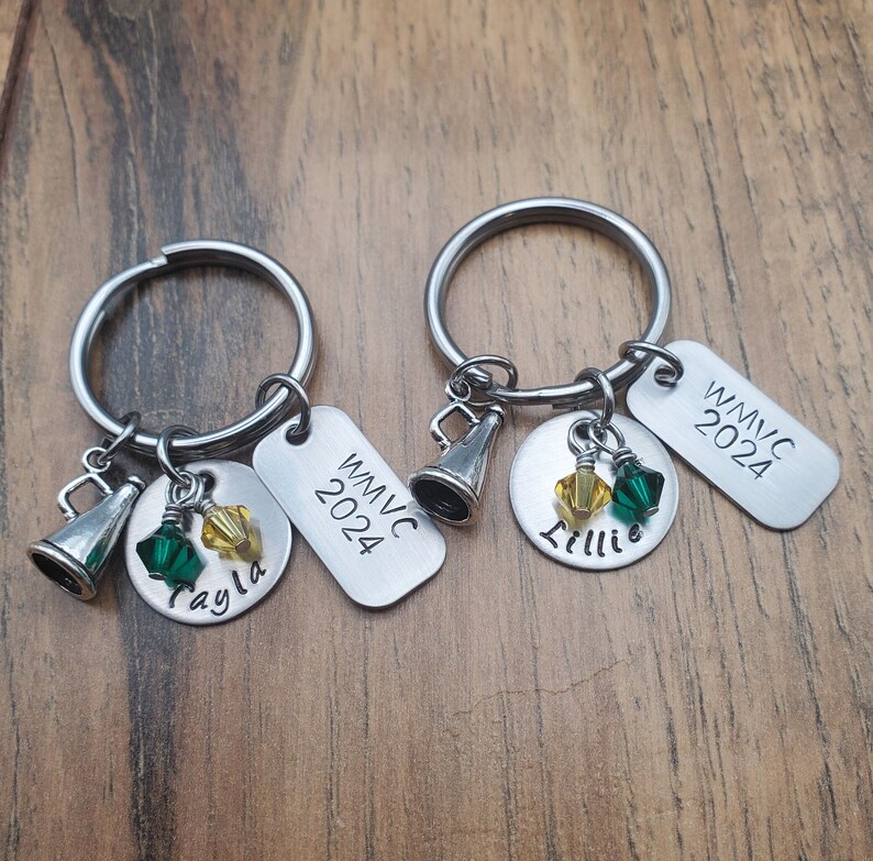 May include: Two silver keychains with charms. Each keychain has a silver tag with the text "WMVC 2024". One keychain has a charm with the name "Tayla" and the other has a charm with the name "Lillie". Both keychains have a silver whistle charm and green and yellow crystal charms.