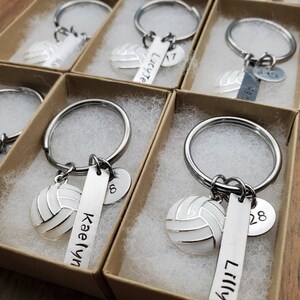 May include: A set of personalized volleyball keychain gifts in small brown boxes. Each keychain features a silver volleyball charm, a silver tag with a name, and a silver charm with a number. The names on the keychains are Kaelyn, Lilly, and Lucy. The numbers on the keychains are 17, 28, and 8.